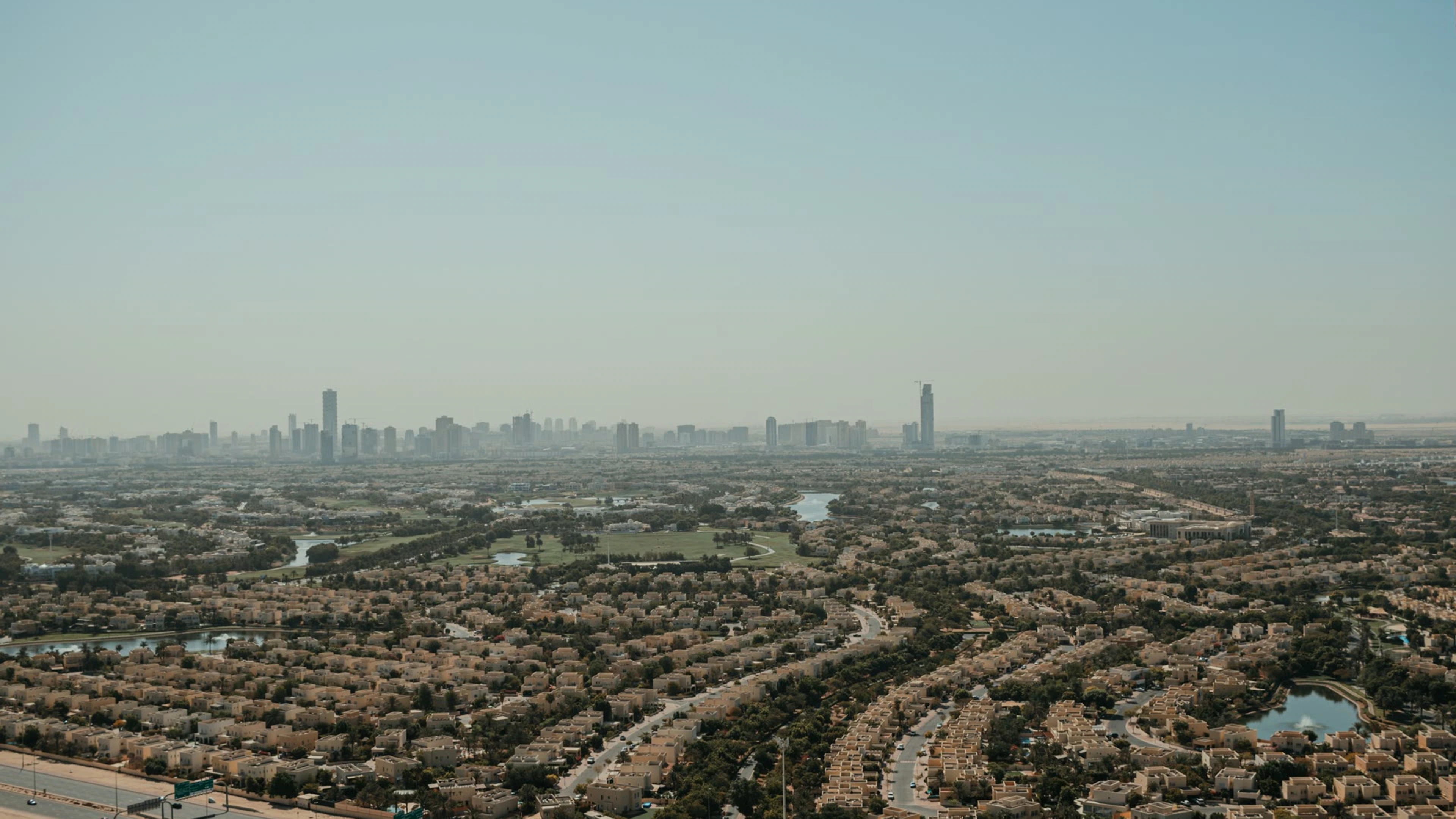 Aerial view of low-rise residential neighbourhoods, lakes and a distant Dubai skyline.